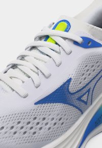 Κοντινό πλάνο λευκού αθλητικού παπουτσιού με μπλε λογότυπο, λευκά κορδόνια, επάνω μέρος από δίχτυ με οπές αερισμού και λεπτομέρεια νέον κίτρινου στη γλώσσα.