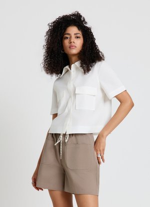 Femme aux cheveux bouclés portant une chemise blanche à manches courtes boutonnée et un short beige à cordon sur un fond uni.