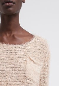 Beige stickad tröja med texterad yta, som har en mjuk pälsliknande lappar och spridda små paljetter, rund halsringning och lös passform.