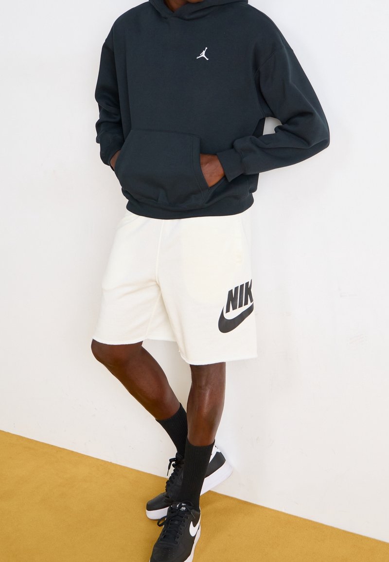 Personne portant un sweat à capuche noir Air Jordan, un short blanc Nike, des chaussettes noires et des baskets noires Nike, debout contre un mur blanc et un sol jaune.