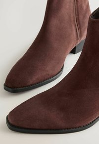 Par de botas de tobillo de ante marrón oscuro con puntas afiladas, suelas negras y cremalleras laterales sobre un fondo claro.