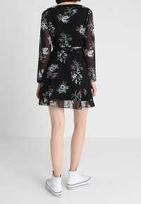 Schwarzes Blumenkleid mit langen, transparenten Ärmeln, tailliertem Schnitt und ausgestelltem Rock. Enthält eine Mischung aus bunten Blumenmustern. Weiße Sneakers sichtbar.
