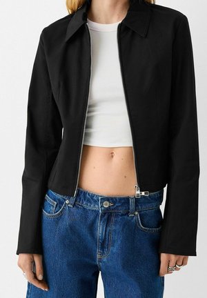 Vrouw draagt een cropped zwarte jas met rits over een witte geribbelde cropped top en een high-waisted blauwe spijkerbroek, waarbij de buik zichtbaar is en ze ringen aan haar linkerhand draagt.