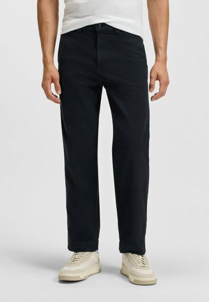 Black cotton trousers with a straight-leg design, featuring a front button closure and side pockets, paired with light-colored sneakers.