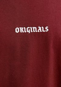 Bordo T-kreklis ar ribotu struktūru, kas satur baltu izšūtu tekstu "ORIGINALS" uzsvarotā fontā. Tuvplāns uz auduma detaļām.