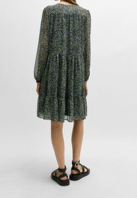 Blümchenmuster-Kleid mit langen, durchsichtigen Ärmeln, geraffter Taille und gestuftem Rock in Dunkelgrün, Blau und Gelb. Kombiniert mit klobigen schwarzen Sandalen.