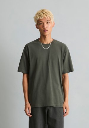Młoda osoba z krótkimi blond włosami, ubrana w luźny ciemnozielony t-shirt, czarne spodnie, srebrny łańcuszek i bransoletkę, stojąca na szarym tle.