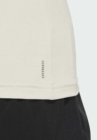 Svetlo bež športna majica z mehkimi teksturami, z malim sivo "AEROREADY" logotipom blizu roba; v kombinaciji z voljo črnimi tkaninami spodaj.