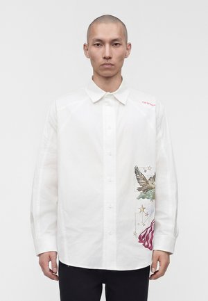 Homme portant une chemise blanche à boutons avec un aigle, des étoiles et des détails rouges abstraits sur le côté droit, debout devant un fond uni.