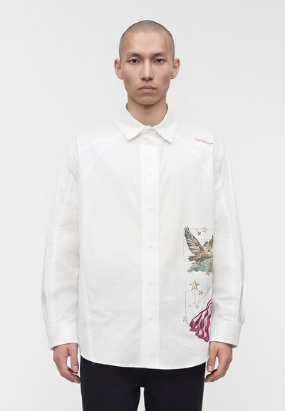 Homme portant une chemise blanche à boutons avec un aigle, des étoiles et des détails rouges abstraits sur le côté droit, debout devant un fond uni.