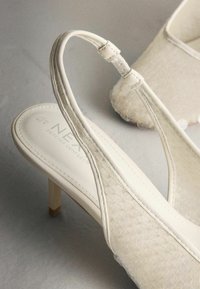 Balti aukštakulniai su dirželiais per kulną turi lygią odinę viršutinę dalį su tekstūruotu paviršiumi, ploną kulną ir subtilų įspaustą logotipą vidinėje pado pusėje.