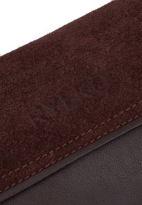 Tessuto in suede marrone con una morbida texture, bordi cuciti, che presenta il logo "RYŁKO" impresso su una sezione in pelle marrone scuro.