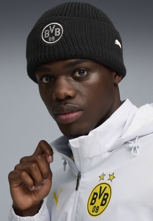 Czarna dzianinowa czapka z okrągłym logo, biała kurtka z zamkiem błyskawicznym i żółtymi akcentami oraz szare tło. Zbliżenie twarzy i dłoni.