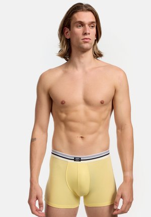 Gelbe Boxershorts aus weichem Stoff, mit einem schwarz-weißen gestreiften Bund, auf dem der Markenname "DIM" deutlich sichtbar ist.