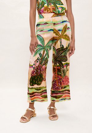 Mujer vistiendo pantalones cortos coloridos con estampados tropicales de palmeras y plantas, combinados con sandalias planas doradas con adornos.