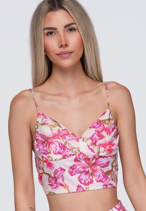 Ung kvinde med langt blond hår iført en hvid crop top med lyserødt blomstertryk og tynde stropper, foran en ensartet lys baggrund.