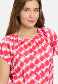 Kortærmet bluse i let materiale med et geometrisk mønster i pink, hvid og rød. Samlet halsudskæring med en lille nøglehulldetalje.