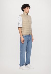 Beige puffer vest med høy krage og glidelås; brukt over en gråmønstret skjorte, kombinert med blå jeans og hvite joggesko.