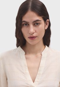 Helle beigefarbene Bluse mit V-Ausschnitt, die ein dezentes Rautenmuster und kontrastierende orangene Nähte aufweist. Weicher Stoff mit glatter Textur.