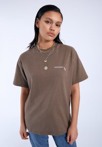 Camiseta de algodón marrón con un corte relajado, que presenta mangas cortas y un logotipo bordado en blanco en el área del lado izquierdo del pecho.