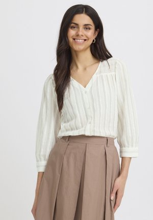 Naeratav naine, kellel on pikad tumedad juuksed, valges triibulises pluusis, millel on volangvarrukad, ja kõrge vöökohaga heledat pruunist volditud seelik.