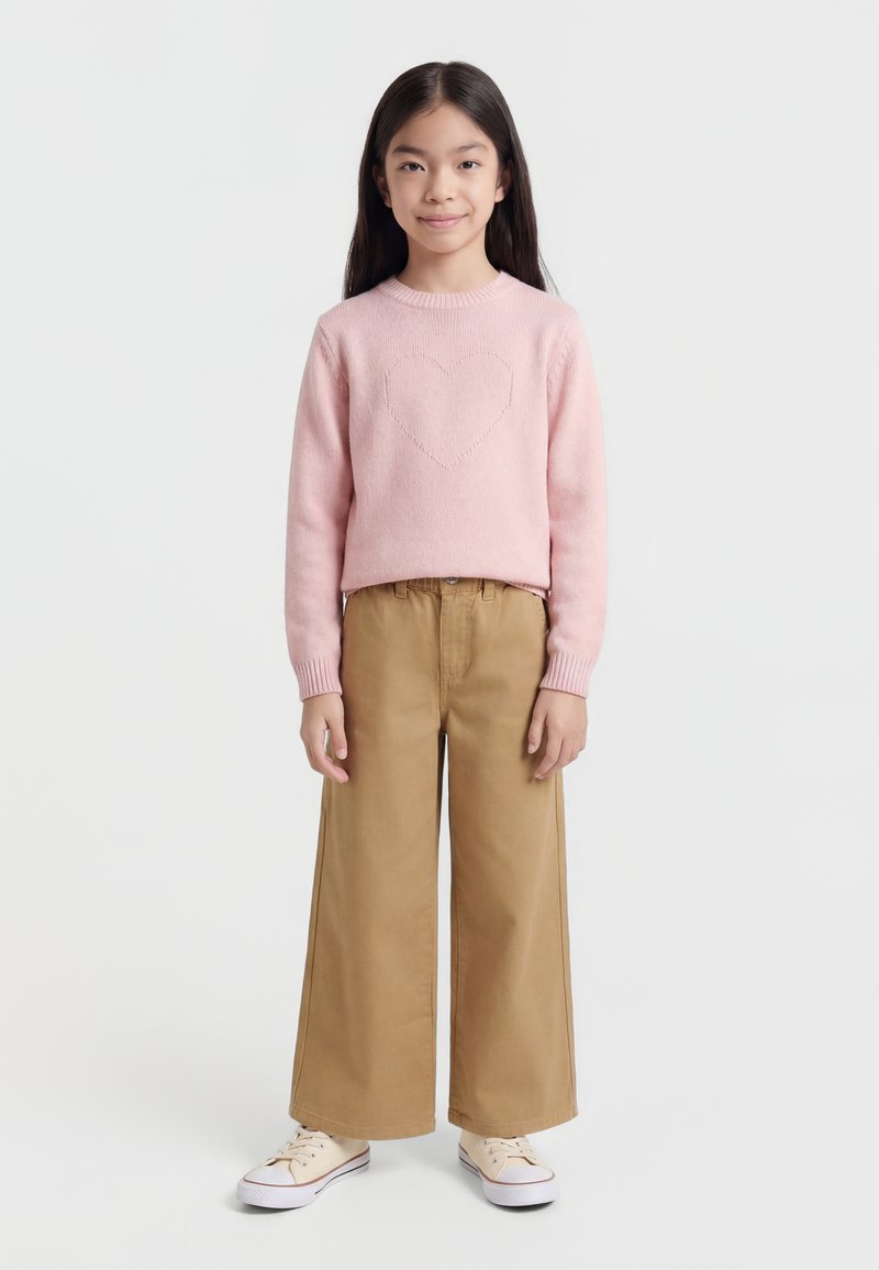 Jeune fille debout, vêtue d'un pull rose avec un détail cœur, d'un pantalon large couleur sable et de baskets beige sur fond blanc.