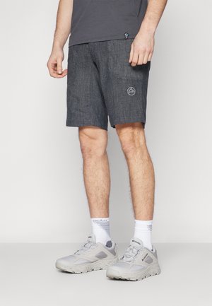 Hombre vistiendo pantalones cortos casuales de color gris oscuro con un logo en la pierna izquierda, calcetines blancos, zapatos deportivos grises y una camiseta gris oscuro.