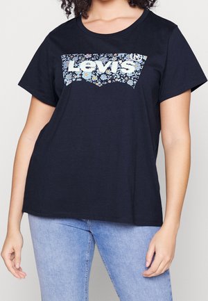 Femme portant un t-shirt Levi's bleu marine avec un logo floral et un jean bleu clair, debout les bras détendus le long du corps.