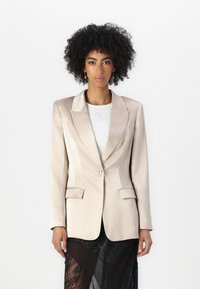 Beige satiini bleiseri, jossa on yksi kultainen nappi, hahlot ja kaksi etutaskua. Päällä valkoinen paita ja musta teksturoitu hame.