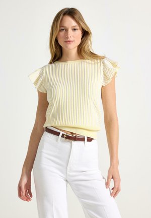 Nainen, joka pukeutuu keltaiseen ribbineulottuun lyhythihaiseen toppiin, jossa on röyhelöhihat, ja valkoisiin korkea vyötäröisiin housuihin, joissa on ruskea vyö, seisoo valkoista taustaa vasten.