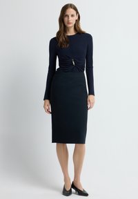 Granatowa bluzka z długim rękawem, z marszczonym przodem i ozdobnym detalem, w połączeniu z dopasowaną, sięgającą kolan spódnicą ołówkową oraz czarnymi szpilkami z cienkim noskiem.