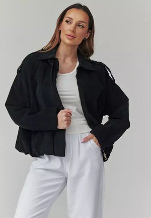 Kobieta w czarnej, oversize'owej kurtce na białym prążkowanym topie i białych spodniach, pozująca z jedną ręką w kieszeni, a drugą na kurtce.