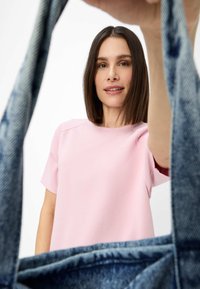 Lyserød kortærmet top med rund hals, der holdes i hånden, med en blå denim totebag i fokus, der har frynsete kanter.