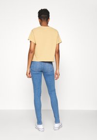 Camiseta beige corta con mangas cortas, combinada con jeans ajustados azules. El modelo lleva zapatillas blancas. El fondo es blanco liso.