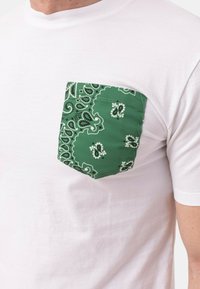 T-shirt bianco con tasca a motivo paisley verde. La tasca è di forma quadrata e presenta disegni intricati in verde scuro e bianco.