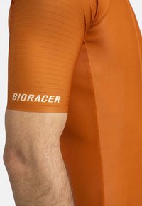Korte mouwen racefietsjersey van gebrand oranje stof met ribbelstructuur. "BIORACER" logo in het wit gedrukt op de mouw.