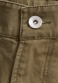 Zoom de una tela caqui de pantalones que presenta un botón de metal con la inscripción "SAIL RACING", costuras visibles y un material de textura suave.