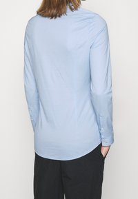 Camicia azzurra chiaro a maniche lunghe in tessuto morbido con orlo curvo, polsini abbottonati e un sottile dettaglio sul colletto. Abbinata a pantaloni neri.