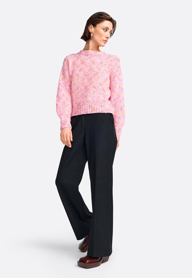 Pull en tricot rose avec un motif texturé, associé à un pantalon ample noir et des chaussures à plateforme marron foncé. Le modèle se tient de profil.
