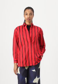 Camicia a righe rosse e nere con chiusura a bottoni, maniche lunghe e colletto a punta. Tessuto morbido; vestibilità casual. Abbinata a pantaloni con motivo.
