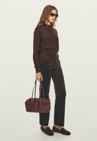 Donna in cardigan bottonato marrone scuro e pantaloni, con una borsa in pelle marrone, che indossa mocassini scuri e occhiali da sole su uno sfondo neutro.
