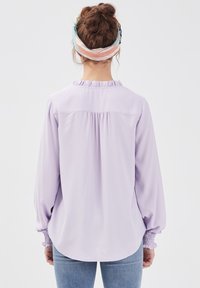 Blusa viola chiaro con scollatura arricciata e polsini elasticizzati. Tessuto morbido, vestibilità leggermente ampia e sottili pieghe sul retro. Abbinata a jeans blu.