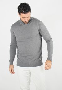 Pull en maille gris au design ajusté, avec des poignets et un ourlet côtelés, et un petit logo sur le bas. Porté sur un pantalon blanc.
