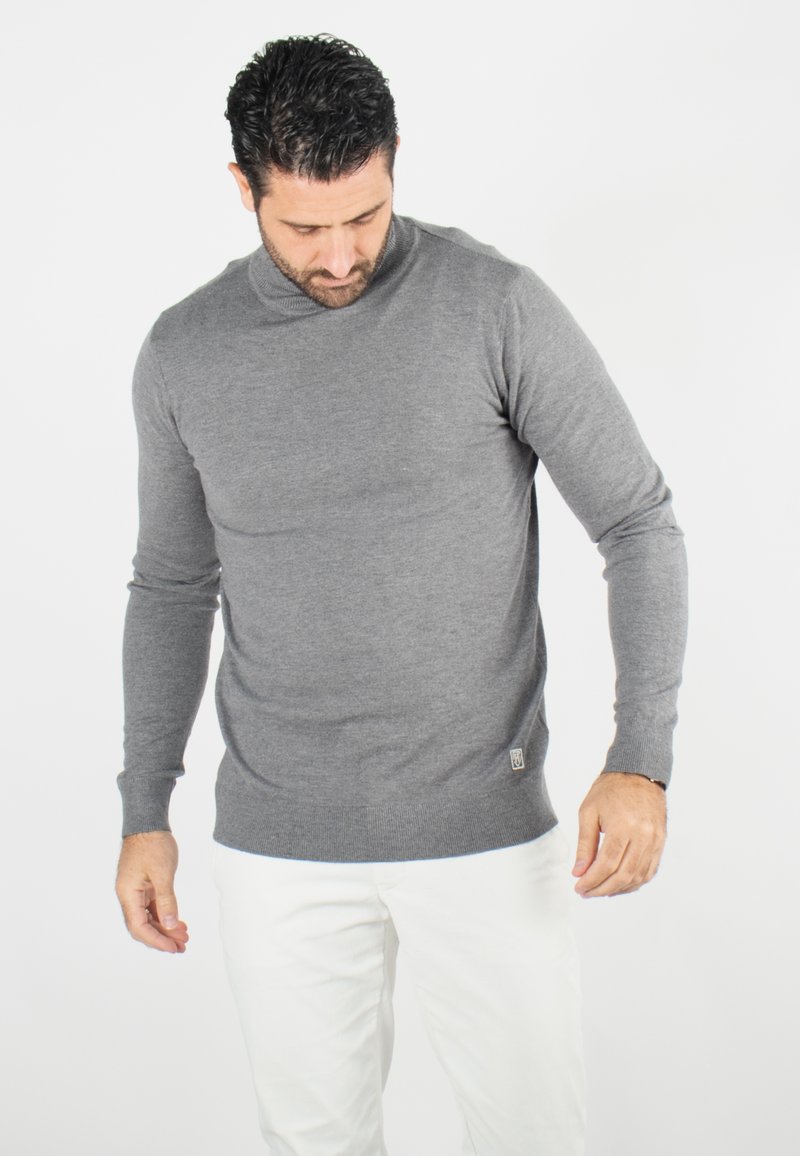Pull en maille gris au design ajusté, avec des poignets et un ourlet côtelés, et un petit logo sur le bas. Porté sur un pantalon blanc.