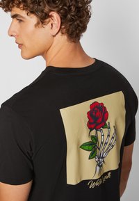 Black t-shirt featuring a large graphic on the back: a red rose held by a skeletal hand against a beige background, with "Wasted Youth" text.