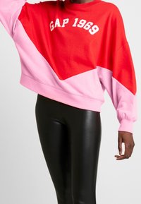 Felpa smanicata rossa e rosa con testo "GAP 1969". Presenta spalle cadute, polsini larghi e un design a blocchi di colore con una texture morbida.