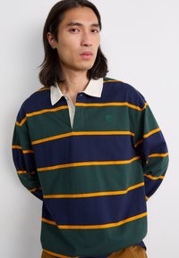 Long-sleeve polo shirt in navy, green, and yellow stripes. Features a cream collar and embroidered logo on the chest. Soft cotton material.