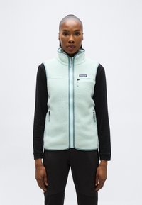 Γυναίκα που φοράει ανοιχτό γαλάζιο φλις γιλέκο με φερμουάρ πάνω από μαύρο μακρυμάνικο πουκάμισο, στέκεται μπροστά σε λευκό απλό φόντο.