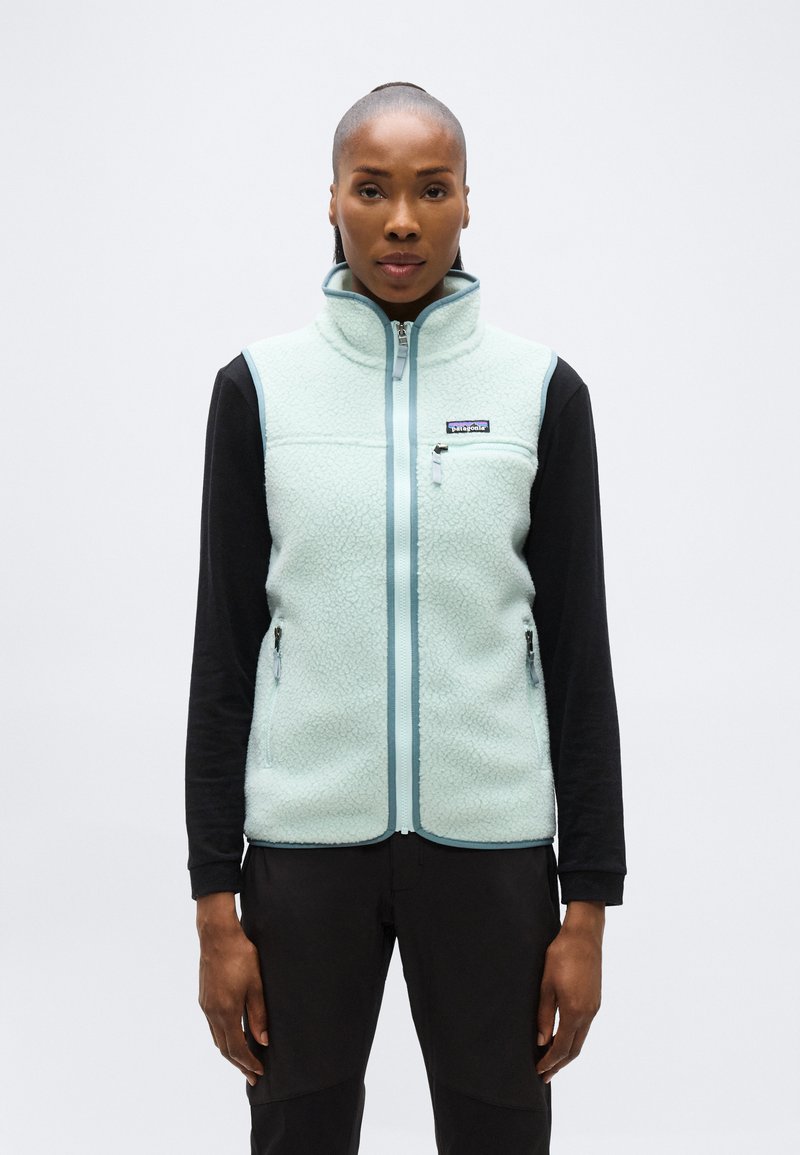 Γυναίκα που φοράει ανοιχτό γαλάζιο φλις γιλέκο με φερμουάρ πάνω από μαύρο μακρυμάνικο πουκάμισο, στέκεται μπροστά σε λευκό απλό φόντο.