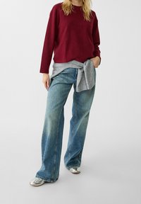 Persoon in een bordeauxrood langof mouwen top, lichtgrijze trui om de taille gebonden, brede blauwe jeans en zilveren schoenen op een simpele witte achtergrond.